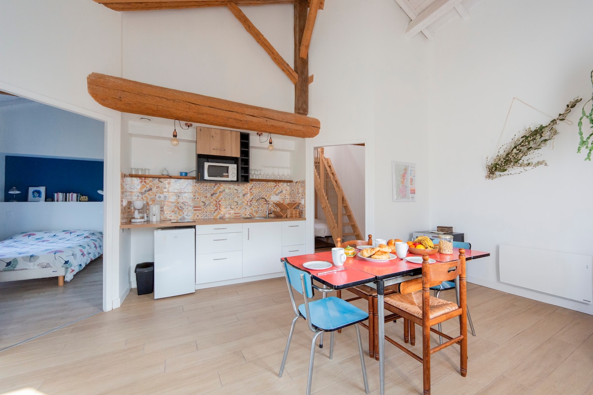 A light-filled kitchen-dining area features a wooden dining table set with breakfast items. Blue and wooden chairs surround the table. The modern kitchen is equipped with cabinets and a microwave, with decorative tiles adding a touch of charm. Exposed beams add height to the space.