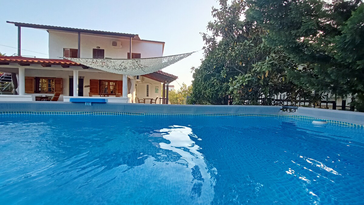A spacious swimming pool is featured in the foreground, reflecting the clear blue sky. The background showcases a two-story house with multiple windows and a shaded area draped with fabric, surrounded by lush greenery.
