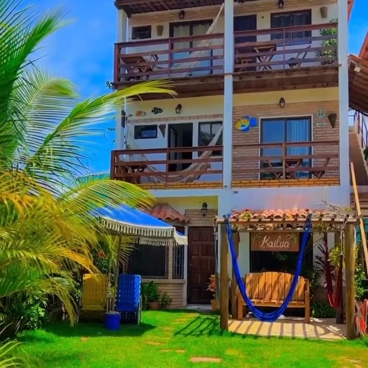 A multi-story building features wooden balconies adorned with decorative elements. Lush greenery surrounds the pathways, while a wooden sign reading 'Kailua' marks the entrance. A hammock hangs nearby, inviting relaxation amid the vibrant atmosphere and clear blue sky.