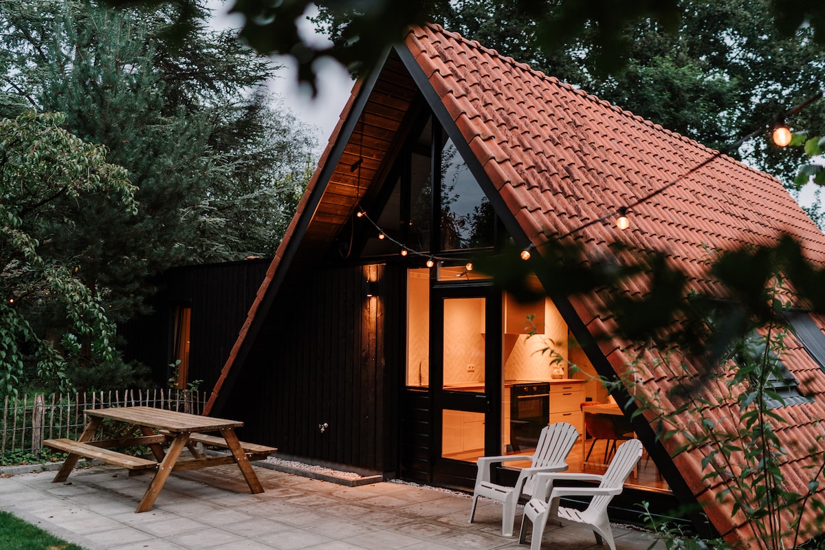 A charming A-frame house features a striking red-tiled roof and large windows that reflect the surrounding greenery. A wooden picnic table is positioned on the patio, complemented by classic white lounge chairs. Soft lighting illuminates the entrance, inviting guests to explore further.