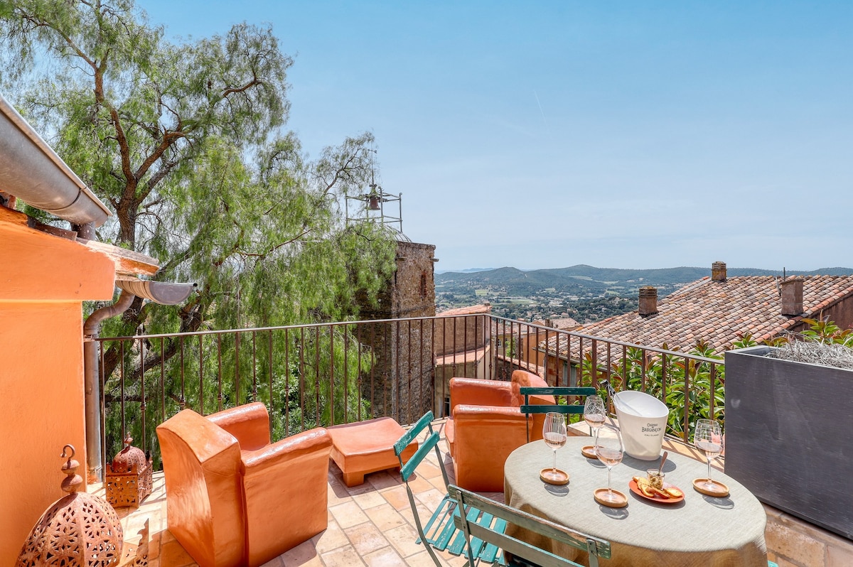 A terrace is presented with panoramic views of the surrounding landscape. Outdoor seating is arranged, including colorful chairs and a circular table. The surface is paved with terracotta tiles, and greenery can be seen nearby, enhancing the space's natural ambiance.