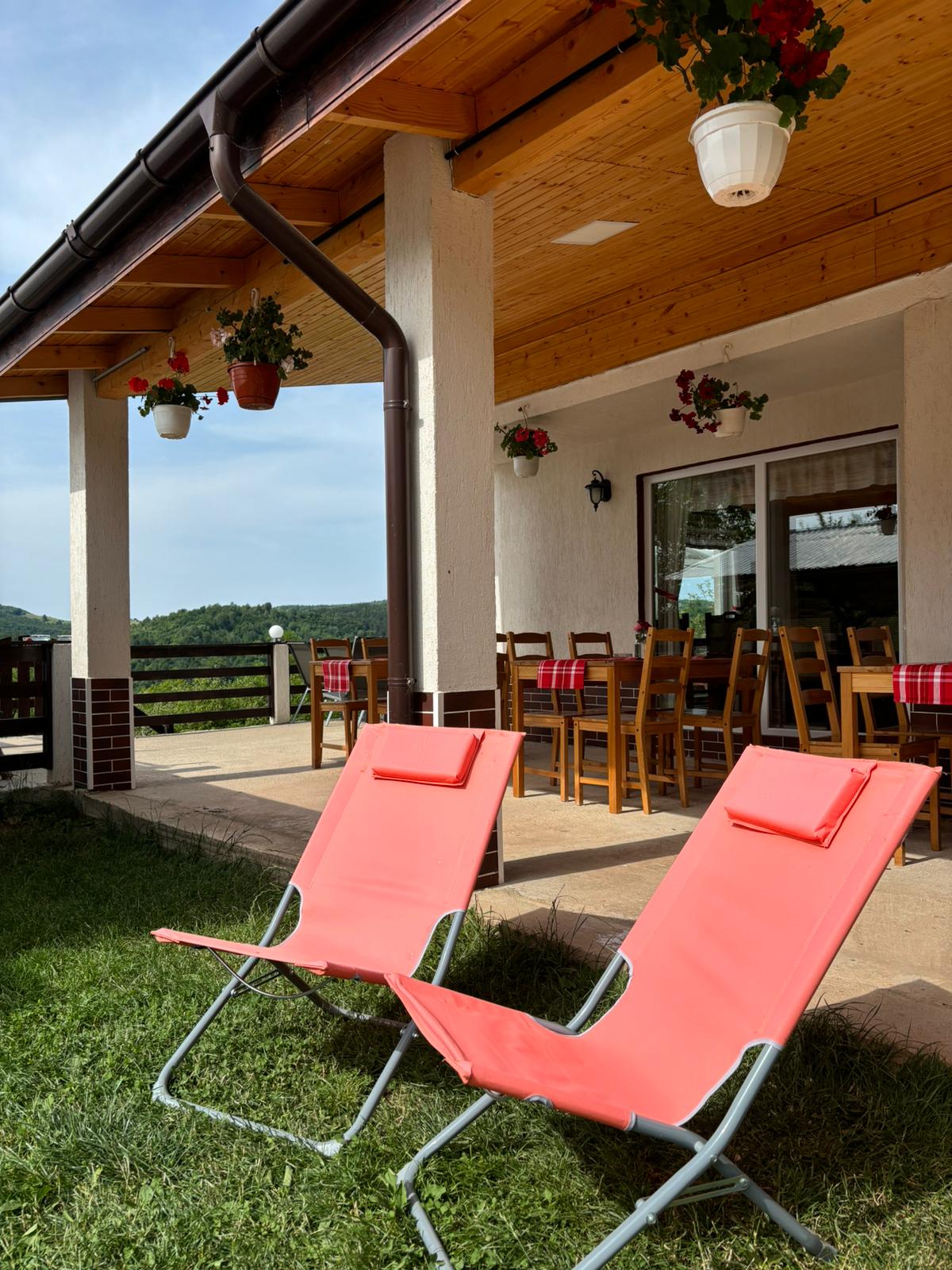 A covered terrace features two bright orange lounge chairs positioned on lush green grass. A wooden dining table with matching chairs is set nearby, with decorative flower pots hanging above, offering a welcoming outdoor dining space with scenic views.