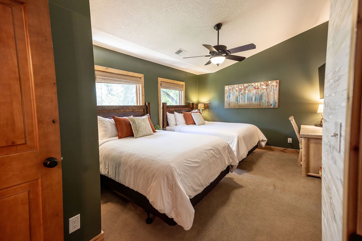 A well-appointed bedroom is featured, with two spacious beds arranged against a green wall. Natural light enters through two windows with wooden frames. A ceiling fan adds comfort, while a bedside table and lamp enhance functionality. A colorful artwork is displayed on the wall.