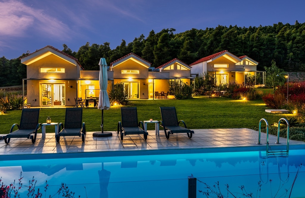 The image displays a serene bungalow complex set against a forested hillside. A sparkling pool reflects the evening sky, surrounded by lounge chairs. Soft outdoor lighting enhances the welcoming ambiance of the spacious grounds, where well-maintained gardens complement the modern architecture.