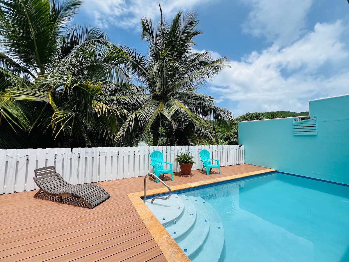 A private patio features a tranquil pool surrounded by wooden decking. Two bright chairs sit beside the pool, complemented by tropical palm trees in the background. The white fence creates a secluded atmosphere, enhancing the inviting setting.