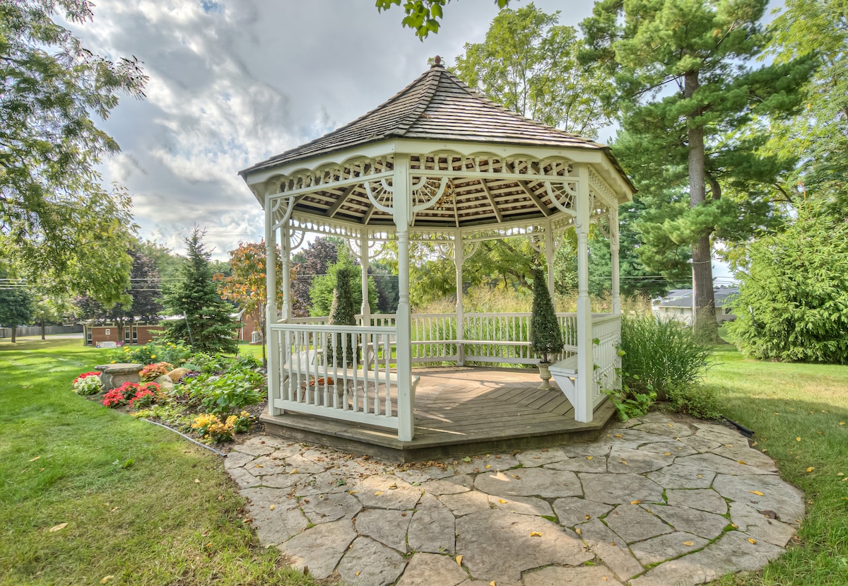 A charming gazebo is set within a landscaped garden, featuring a circular stone patio surrounded by flower beds. Lush greenery frames the structure, with trees providing a natural backdrop. The gazebo's wooden framework showcases intricate design details, inviting relaxation in the tranquil outdoor space.