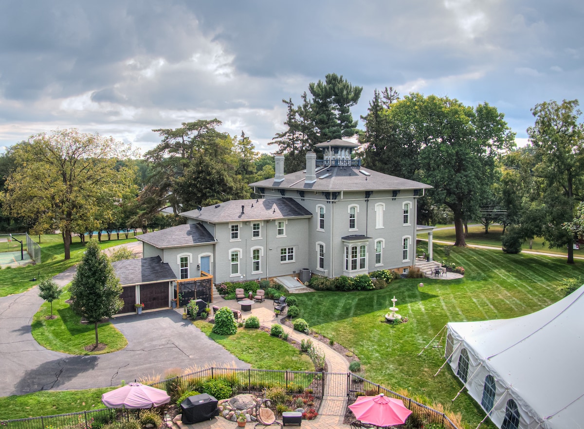 The villa's exterior is captured, showcasing a historic Italianate style with a gray facade. Lush green lawns surround the property, dotted with trees. A spacious patio area features seating, and a nearby tent is set up for events. The tennis court is visible in the background.