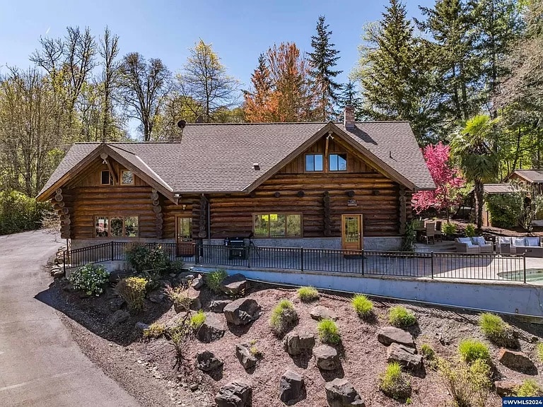A rustic log home is framed by lush landscaping. Large windows offer views of the surrounding trees, while a circular driveway enhances accessibility. The combination of natural stone and greenery complements the home's wooden exterior, showcasing a harmonious blend of nature and architecture.