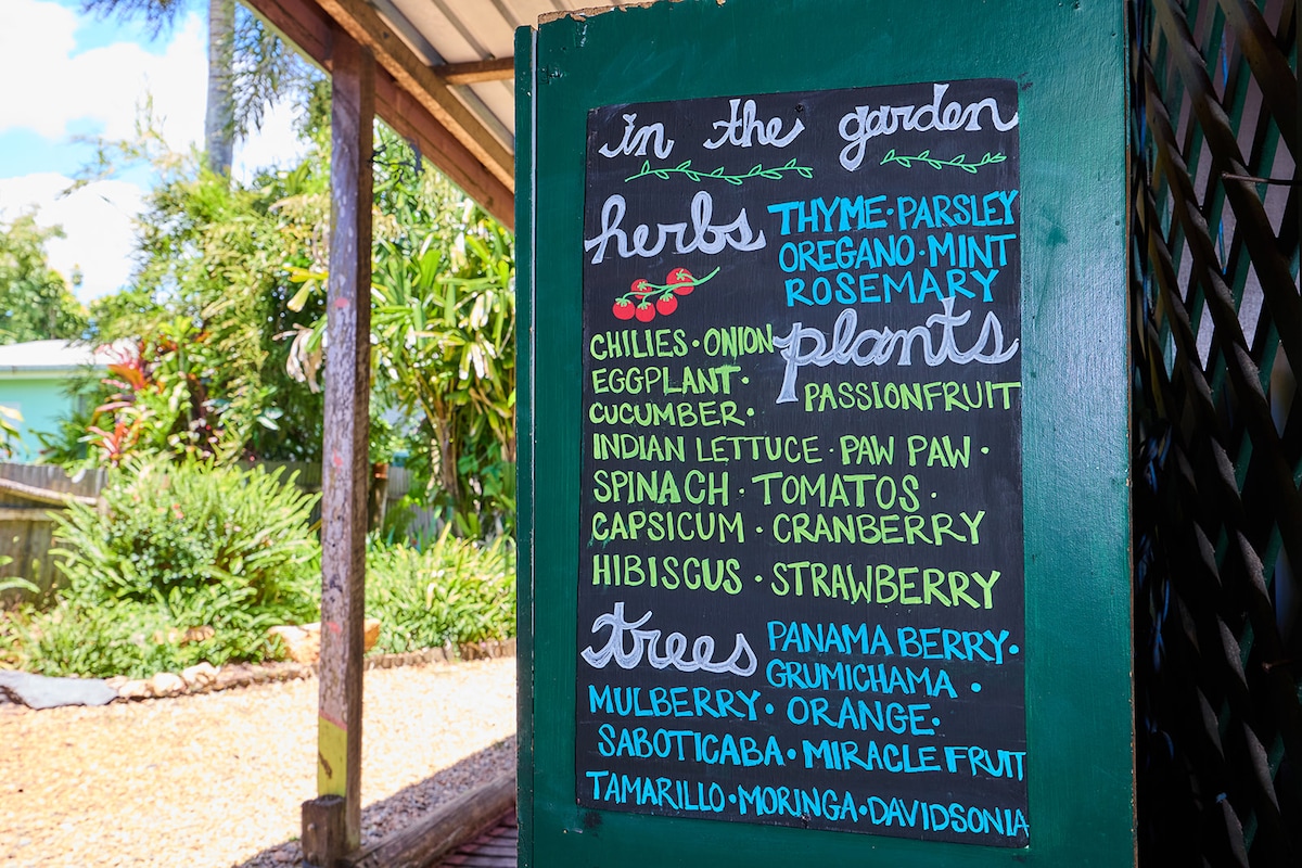 A colorful chalkboard displays a variety of herbs and plants available in the garden. Listed items include thyme, parsley, rosemary, and various vegetables and fruits, such as spinach and strawberries, against a backdrop of lush green foliage.