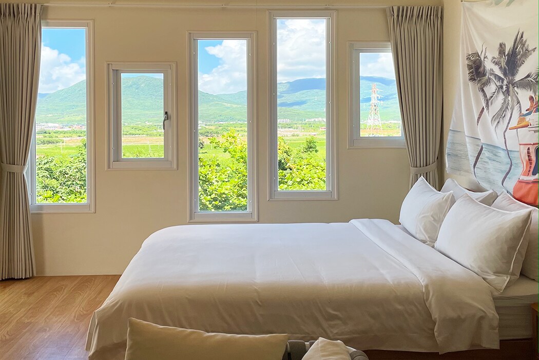 A bright and airy bedroom features a spacious bed with crisp white linens. Large windows provide scenic views of rolling hills and greenery, allowing ample natural light to fill the room. Soft curtains frame the windows, enhancing the serene atmosphere.