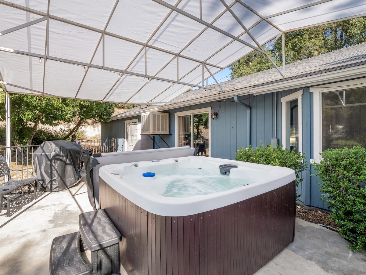 A spacious hot tub is situated on a covered patio, surrounded by greenery. Nearby, a propane BBQ grill and outdoor seating are visible, providing ample space for relaxation. The exterior of the home displays a blue siding, with multiple windows allowing natural light.