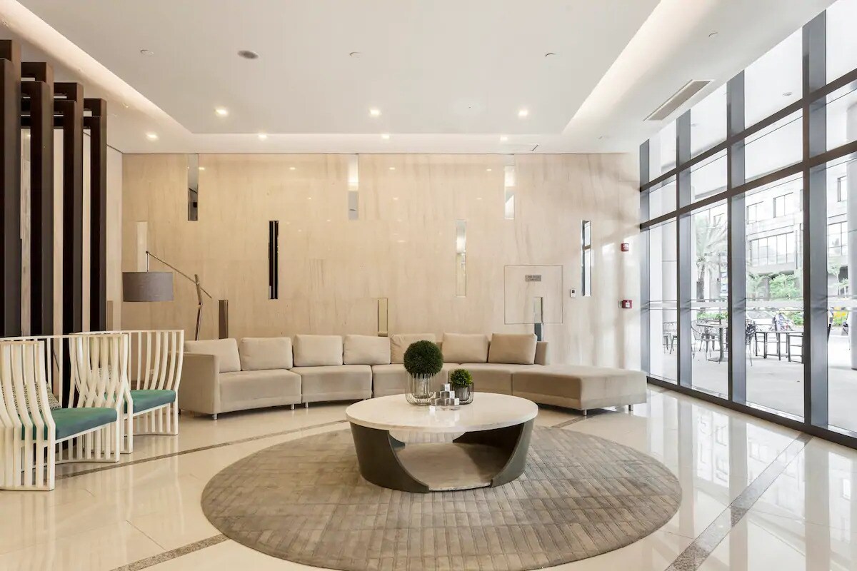 The lobby area features a modern design with a marble floor and large glass windows allowing natural light. A circular area rug is positioned beneath a low coffee table, surrounded by a spacious sectional sofa and unique seating arrangements.