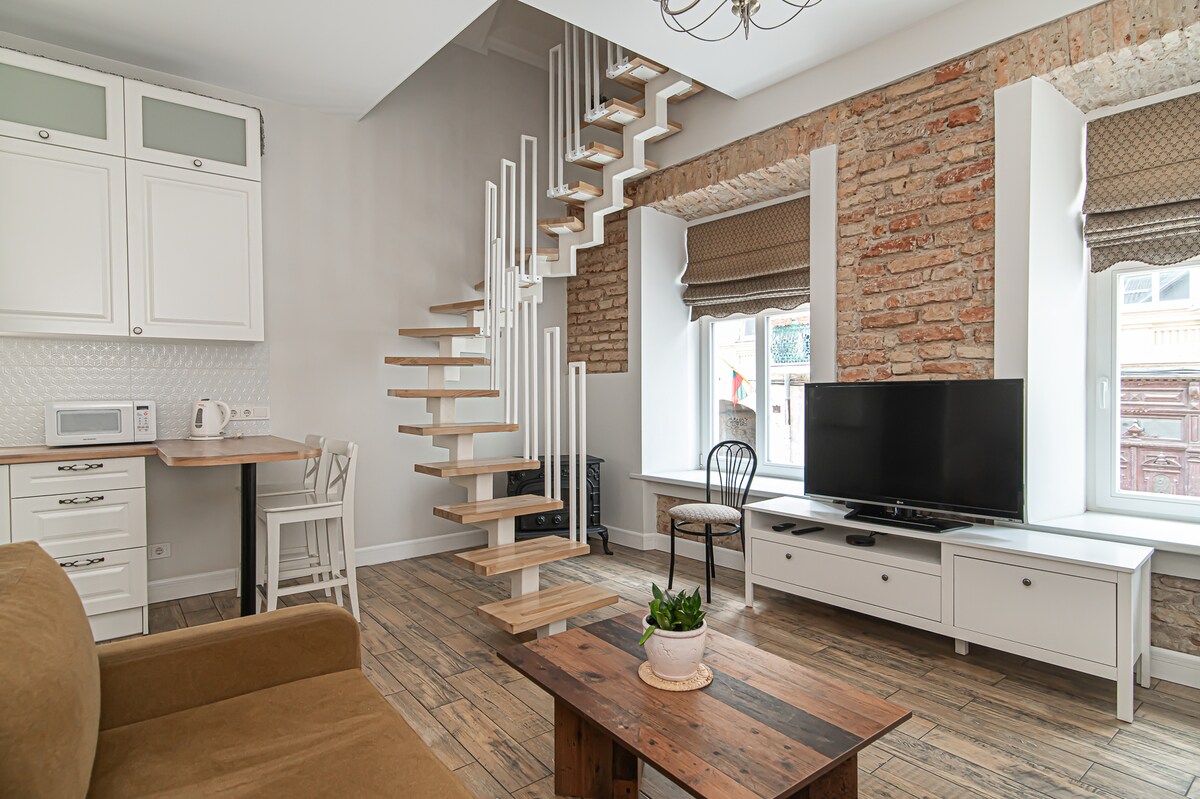 A modern living space features a light-colored sofa and a wooden coffee table. A staircase leads to the upper floor, and a television is displayed on a white cabinet. Bright windows with soft coverings provide natural light, while exposed brick walls add character.