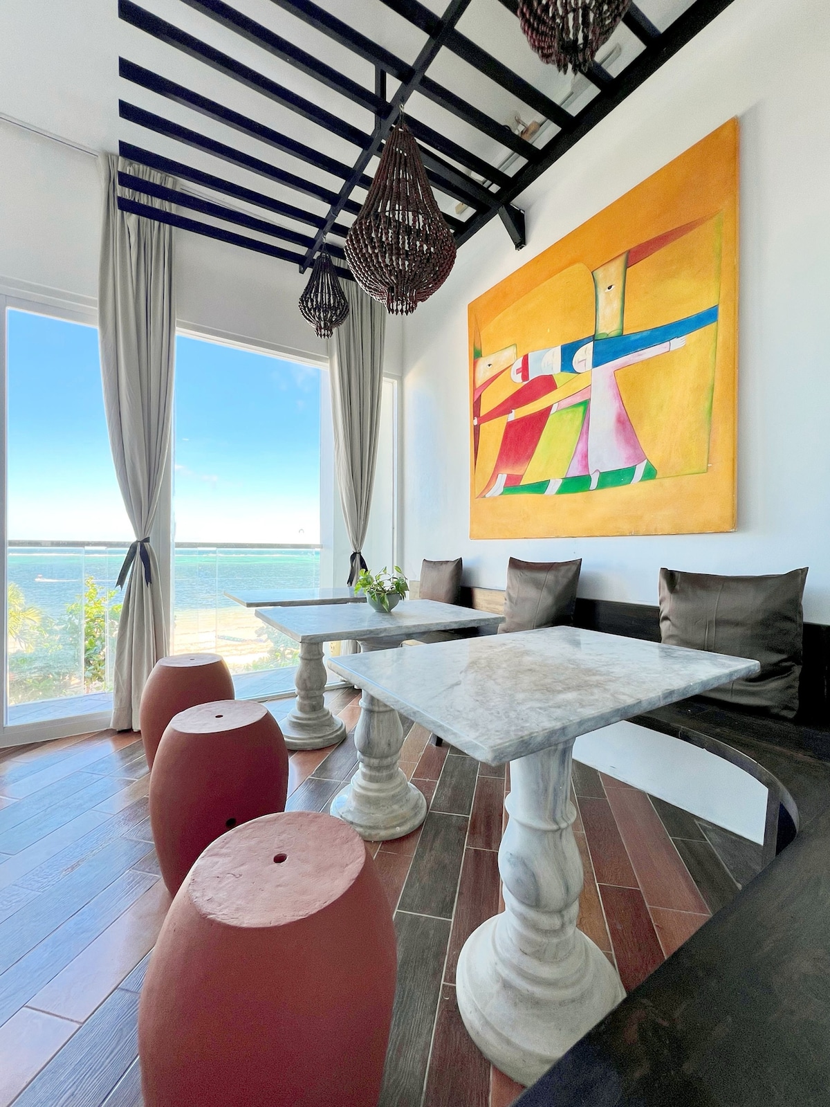 A bright seating area features a marble table surrounded by unique stools. Large windows provide a full view of the ocean, while art adorns the wall, contributing to a colorful and relaxing atmosphere. Soft curtains frame the space, adding a touch of elegance.