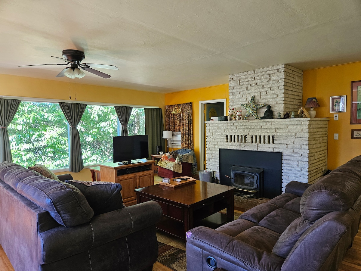 A spacious living room is characterized by two large, comfortable sofas and a wooden coffee table. A fireplace with a white brick surround serves as a focal point, while large windows offer views of the surrounding greenery. A television is positioned nearby.