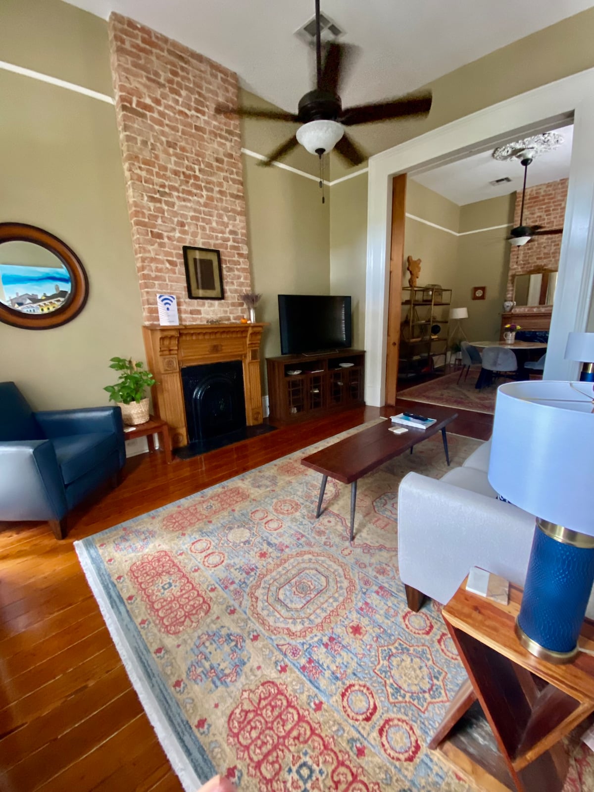 A cozy living area features exposed brick walls and warm hardwood flooring. A plush area rug adds texture beneath a mid-century coffee table. A 56-inch TV is positioned near a decorative fireplace, with comfortable seating provided by an armchair and a modern lamp illuminating the space.