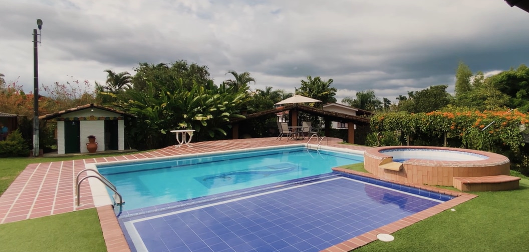 An outdoor pool area features a large swimming pool alongside a smaller hot tub, both surrounded by lush greenery. Lounge chairs are positioned nearby under a canopy, providing shaded seating options. The space is designed for relaxation and recreational activities.
