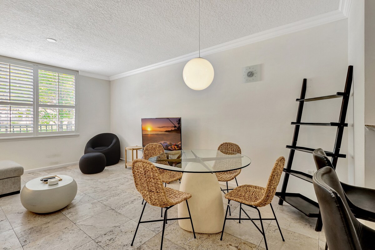 A bright living area features a round glass dining table surrounded by four woven chairs. A comfortable black armchair and a light gray ottoman provide additional seating. Natural light filters through the window, enhancing the space along with a modern television and decorative ladder shelf.
