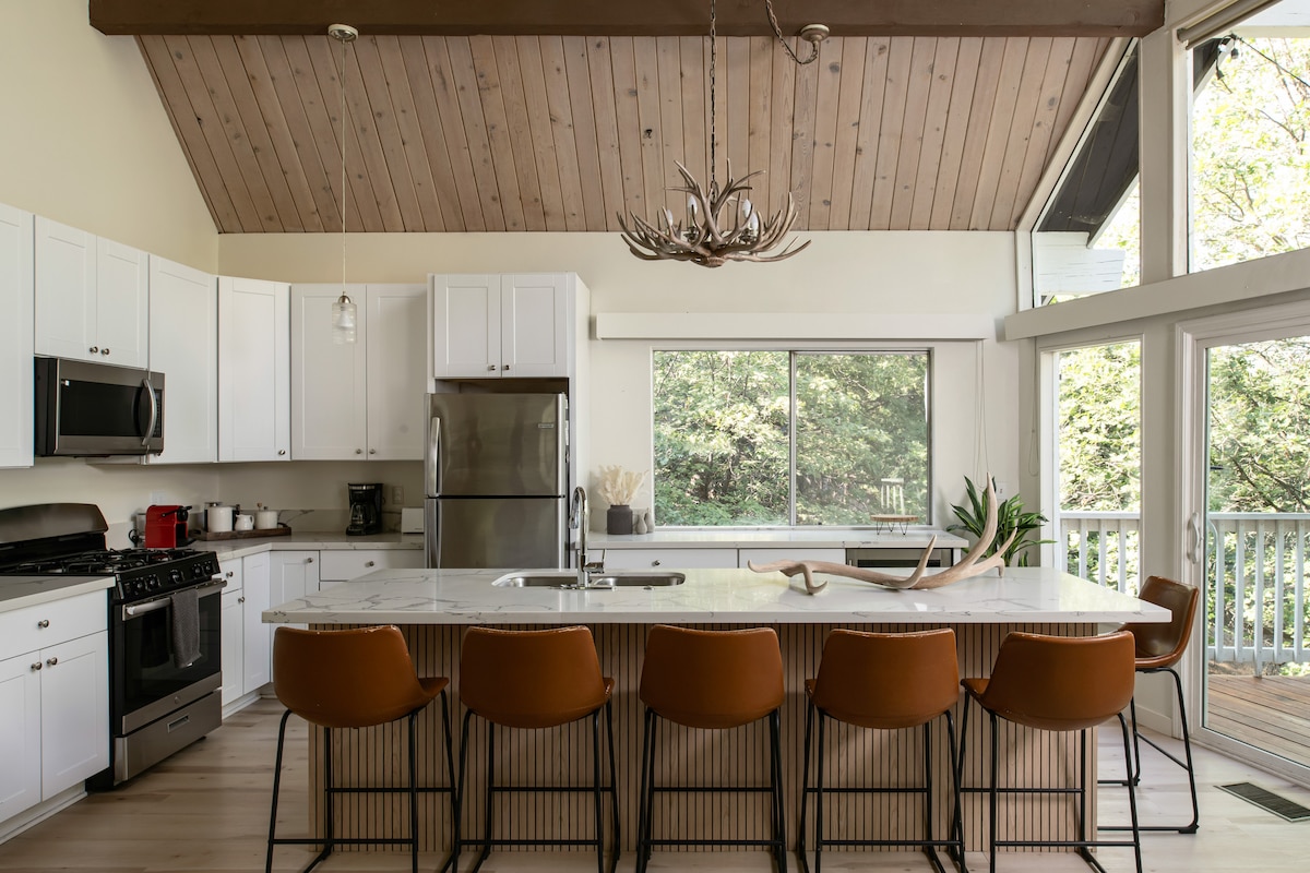 The kitchen features a spacious island with seating for eight, complemented by modern appliances and white cabinetry. Large windows allow natural light to fill the room, while the wooden ceiling adds warmth to the open-concept space. A view of greenery is visible through the glass doors.