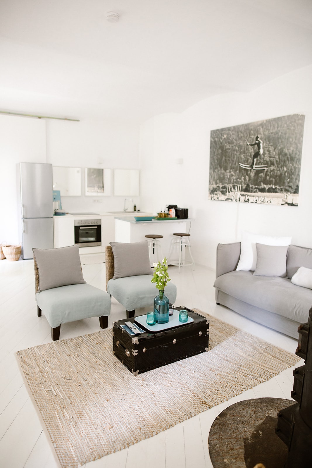 A bright living area features a modern sofa and two armchairs, arranged around a rustic coffee table. The white walls and light wood floors enhance the space's openness. A well-equipped kitchen is visible in the background, complemented by a large refrigerator and contemporary artwork.