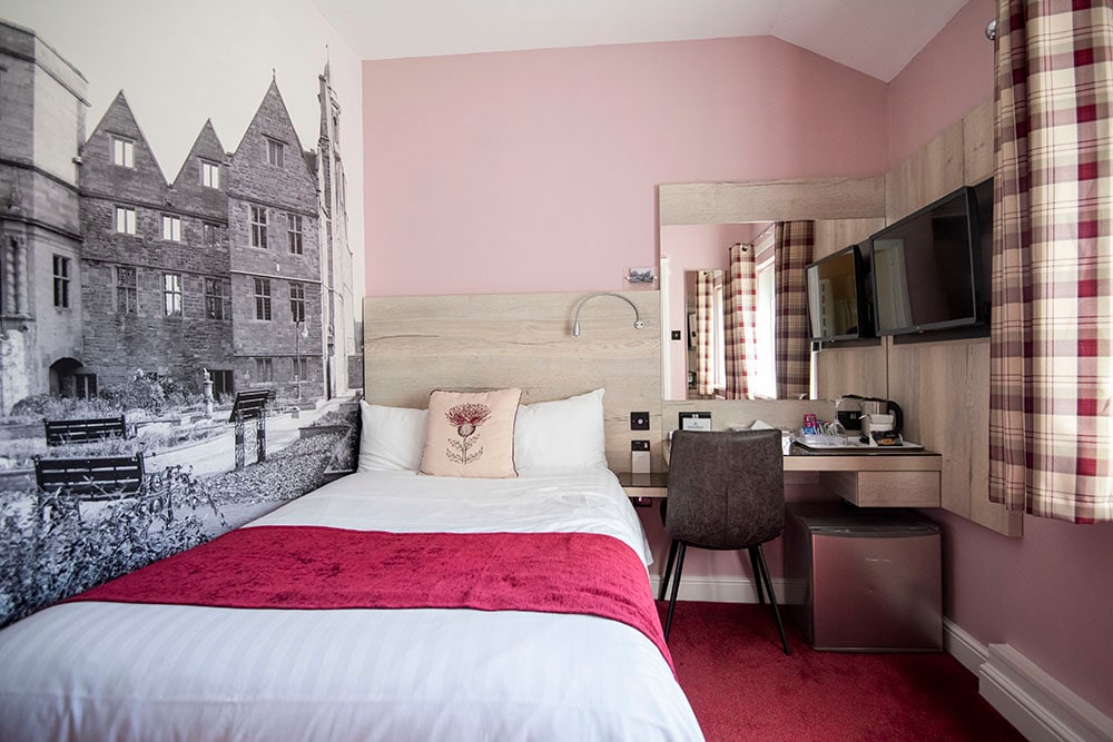 A single room is presented with a compact layout, featuring a spacious bed with a red accent blanket. An artistic black-and-white mural of an old building adorns one wall. A mirrored desk with a chair, a smart TV, and a mini fridge are also included.