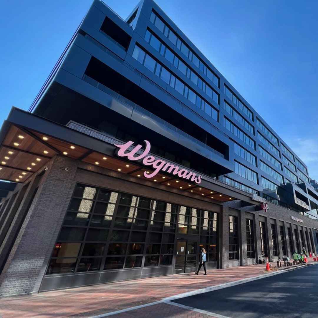 A modern Wegmans grocery store is featured in the image, showcasing a large, prominent sign and expansive glass windows. The building has a sleek design with multiple stories, set against a bright blue sky. The surrounding area includes pathways and construction barriers.