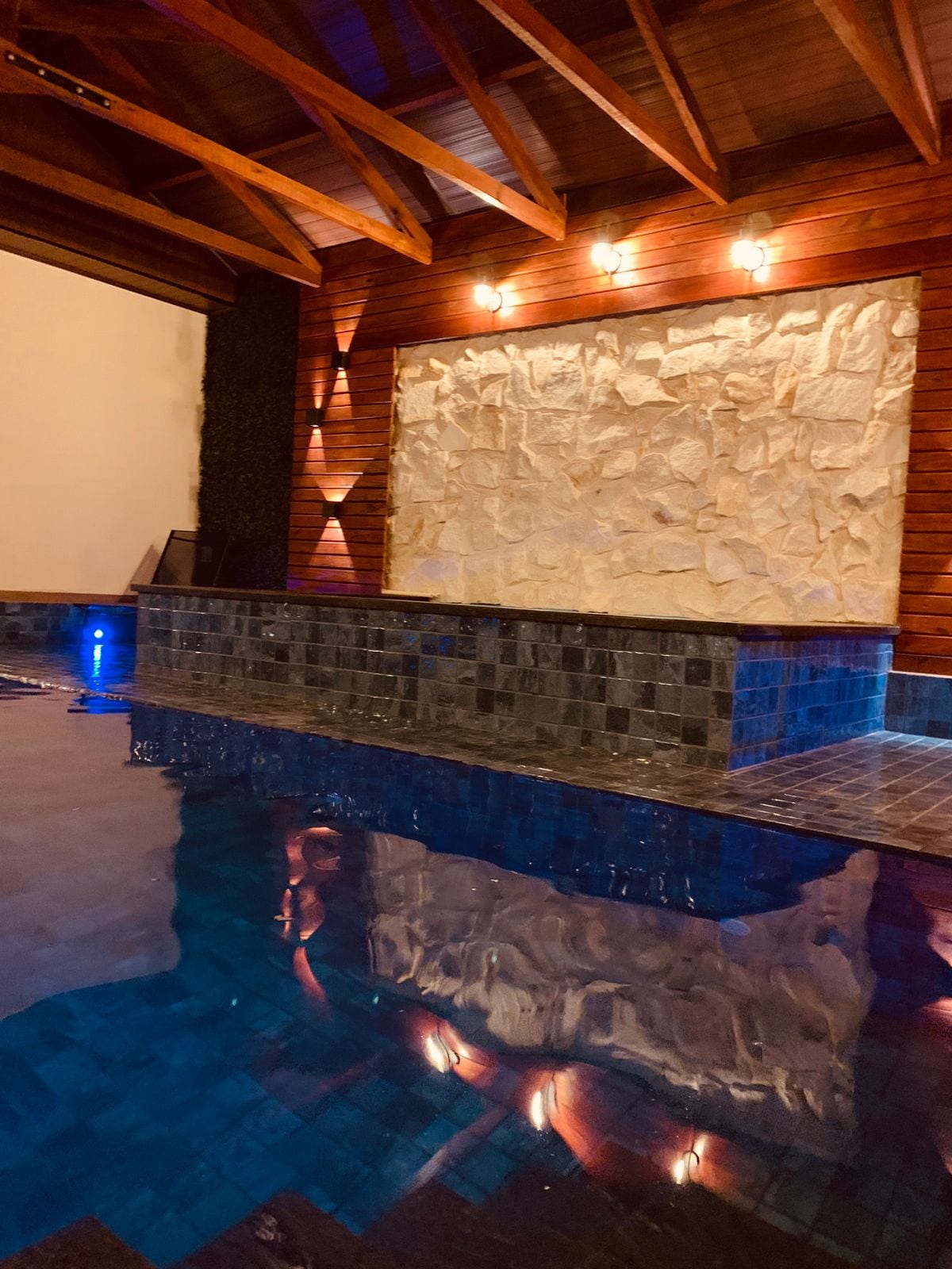 An indoor pool is captured, featuring a smooth stone wall illuminated by soft lighting. The water reflects the architectural elements and the wooden ceiling above, enhancing the tranquil environment.