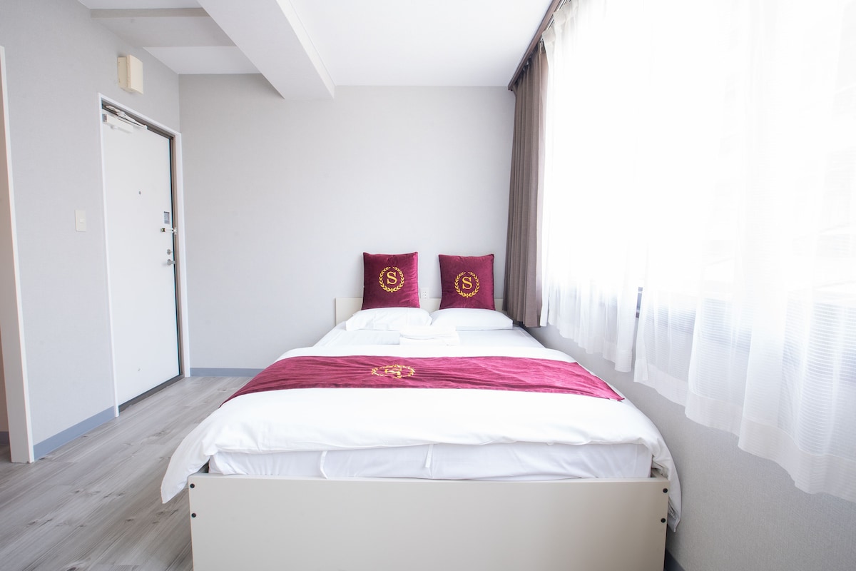 A spacious bed is positioned against a light grey wall, complemented by two decorative pillows. Natural light filters in through sheer curtains, illuminating the room's neutral tones. A clear entry door is visible, enhancing the inviting atmosphere of the space.