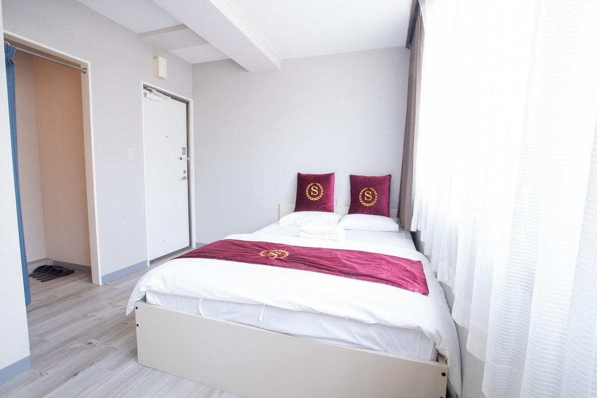 A bright sleeping area features a spacious bed adorned with burgundy accent pillows. Natural light fills the room through large windows, which are partially covered by sheer white curtains. A minimalist design is highlighted by the light wood flooring and clean, neutral walls.