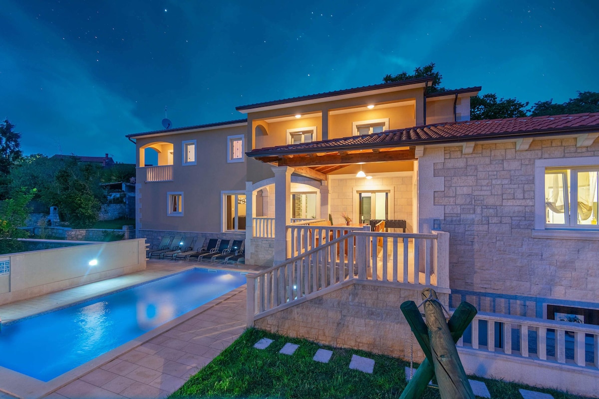 The villa is depicted in the evening light, showcasing a spacious exterior with a pool surrounded by lounge chairs. The covered terrace features a wooden ceiling, and multiple large windows illuminate the rooms within, emphasizing the property’s inviting ambiance against a clear night sky.