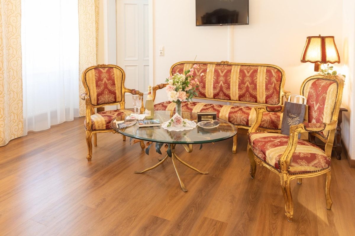 A refined living area features an ornate gold-framed sofa and matching chairs with rich red upholstery. A glass coffee table is centered among the seating, adorned with a floral arrangement and decorative items. Natural light filters through sheer curtains, enhancing the room's elegant ambiance.
