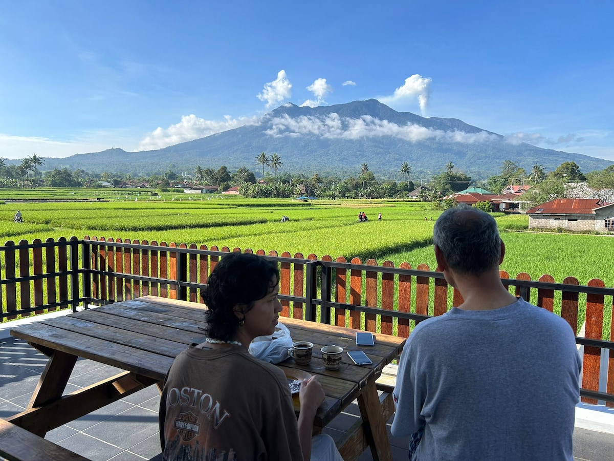 A wooden table sits on a terrace, with two guests enjoying their time while facing a lush green landscape of rice fields. In the background, Mount Marapi rises, framed by a blue sky and scattered clouds, creating a serene outdoor setting.