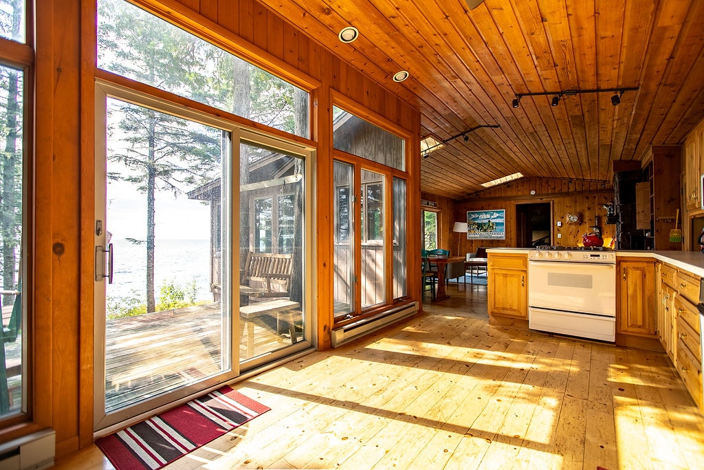 An open-concept living area features warm wood paneling and large windows that fill the space with natural light. The kitchen is equipped with wooden cabinetry and modern appliances. A cozy seating area with a view of the lake is available, enhancing the connection to nature.