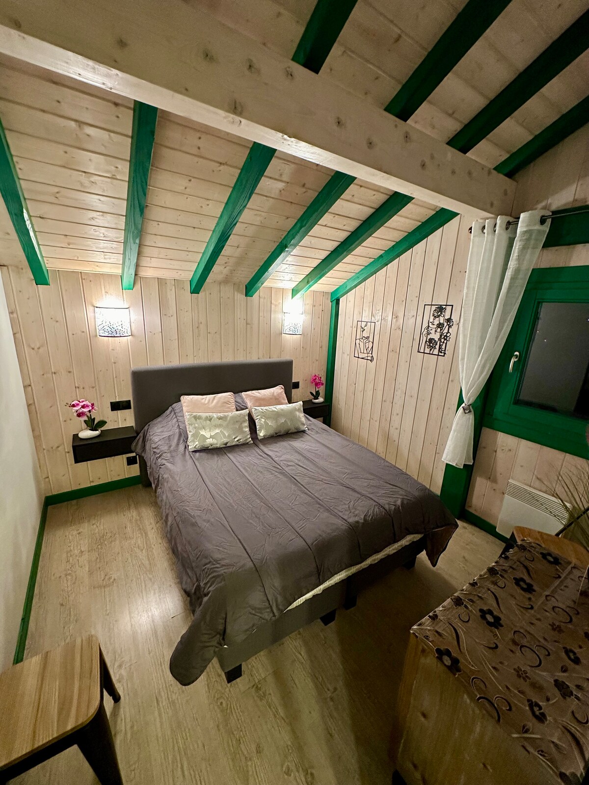A cozy bedroom is presented with wooden paneling and a sloped ceiling. A spacious bed is centered in the room, adorned with light-colored pillows. Soft lighting from wall sconces creates a calm ambiance. A window offers views of the outside, framed by light curtains.