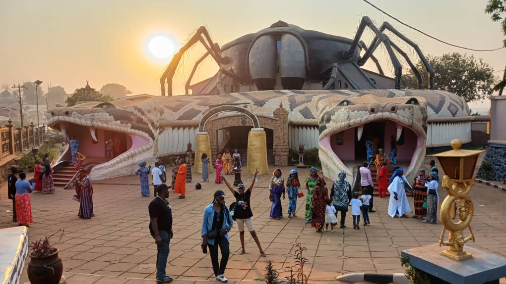 A unique architectural structure resembling a snake and a spider is displayed under a rising sun. Visitors are seen exploring the site, which features distinct entrances shaped like animal heads. The surrounding area is paved, with trees visible in the background.