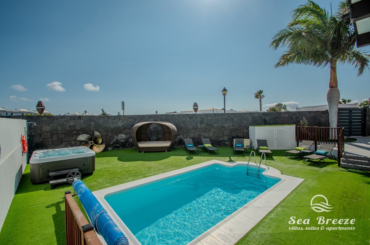 A private outdoor space features a heated swimming pool alongside a jacuzzi. Comfortable loungers are positioned on vibrant green grass, and a shaded seating area offers a relaxing spot. Palm trees sway in the gentle breeze under a clear blue sky.