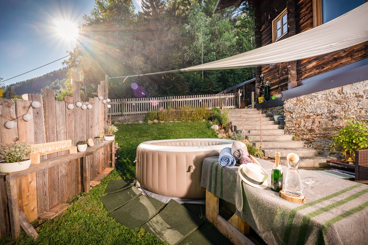 An outdoor space features a private hot tub surrounded by greenery. Wooden fencing adds privacy, and a small table with a blanket and drinks is placed nearby. Stairs lead to the entrance of a rustic wooden building, with sunlight streaming in from a clear sky.