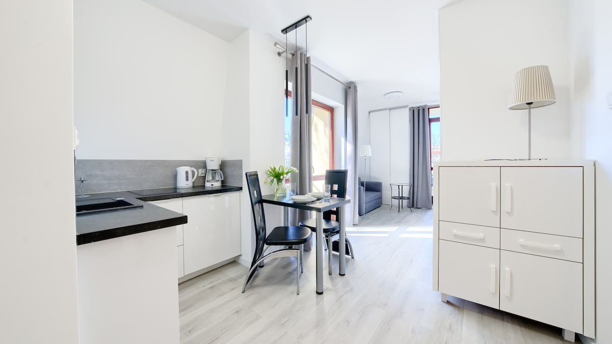 A modern studio apartment features a fully equipped kitchenette and a dining area with a small table and black chairs. Natural light enhances the open space, where a gray sofa is visible in the background, alongside a light-filled area with windows and subtle decor.