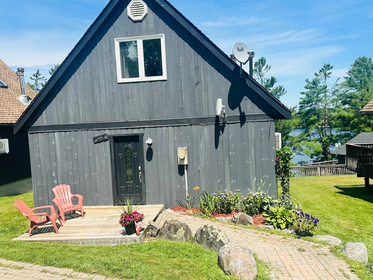 A charming A-frame cottage features a dark wood exterior and a welcoming entrance, flanked by vibrant flowerbeds. Two Adirondack chairs are positioned on a small deck, while a flagstone path leads through a manicured lawn to a scenic backdrop of trees and water.