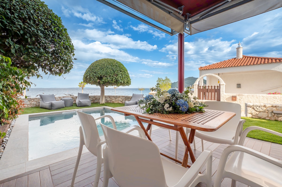 An outdoor dining space is situated alongside a refreshing pool, surrounded by plush lounge chairs. Lush greenery frames the area, with the sea visible in the background. A wooden table is set with floral arrangements, all beneath a shaded awning that enhances the inviting atmosphere.