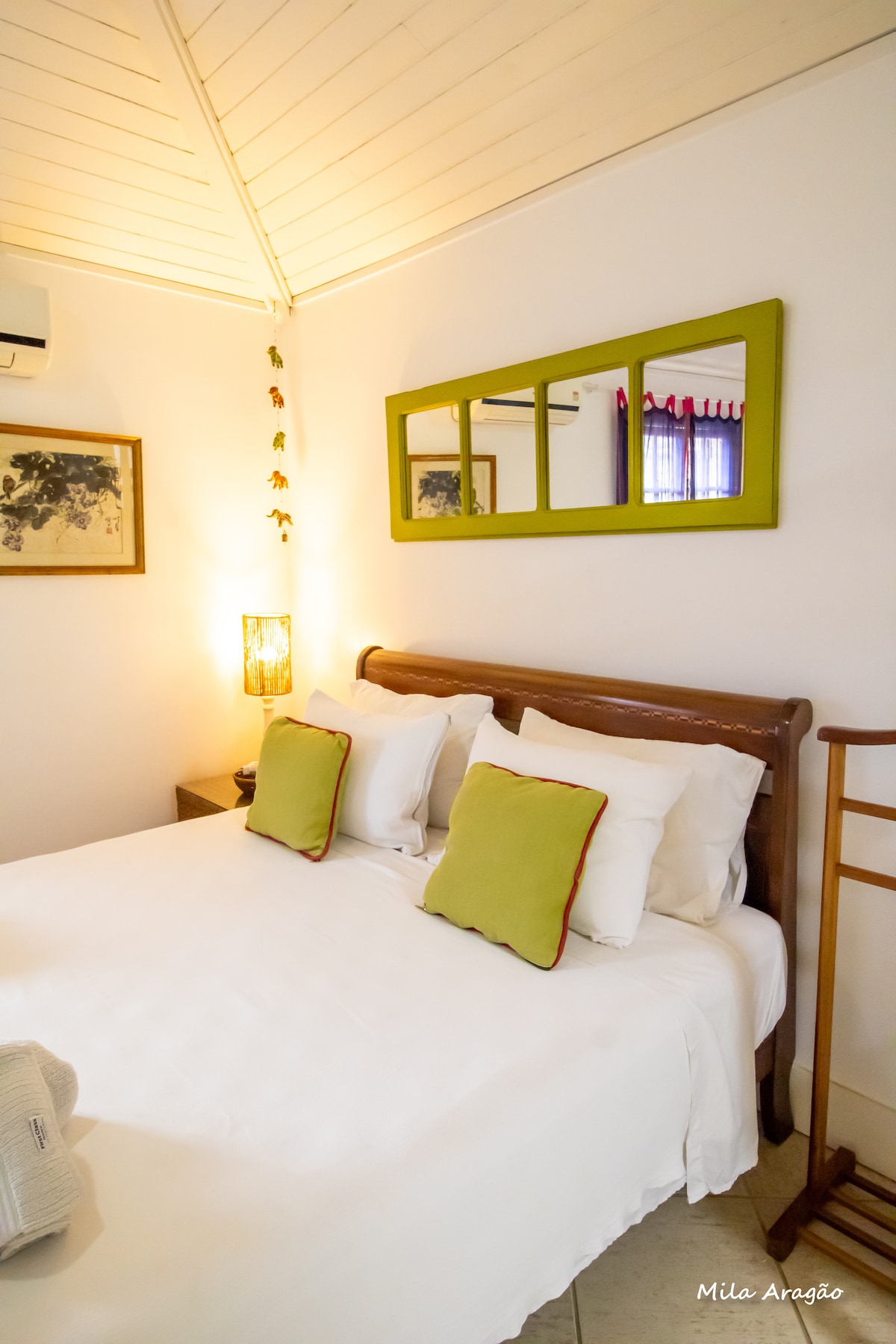 A cozy sleeping area is featured, with a spacious bed adorned with white linens and two green accent pillows. A wooden mirror with a green frame adds a decorative touch, while light filters through a window dressed with burgundy curtains.