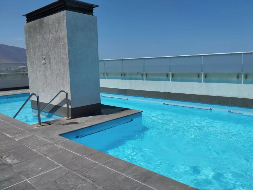 A rooftop pool is visible, surrounded by a tiled deck. The water is clear and blue, with a contrasting light-colored structure featuring a chimney at one end. The expansive view of the sky and mountains contributes to a serene atmosphere.