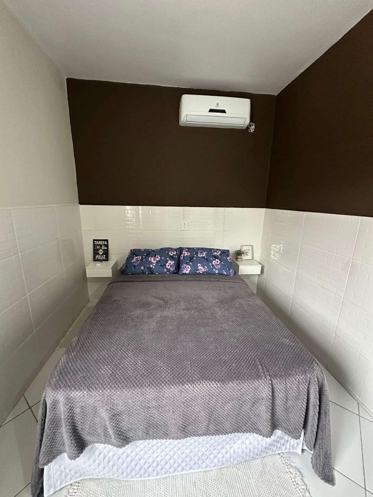 A cozy sleeping area features a spacious bed adorned with decorative pillows and a soft blanket. Air conditioning is installed above, and simple bedside tables are positioned on either side, complemented by minimalist decor on the walls.