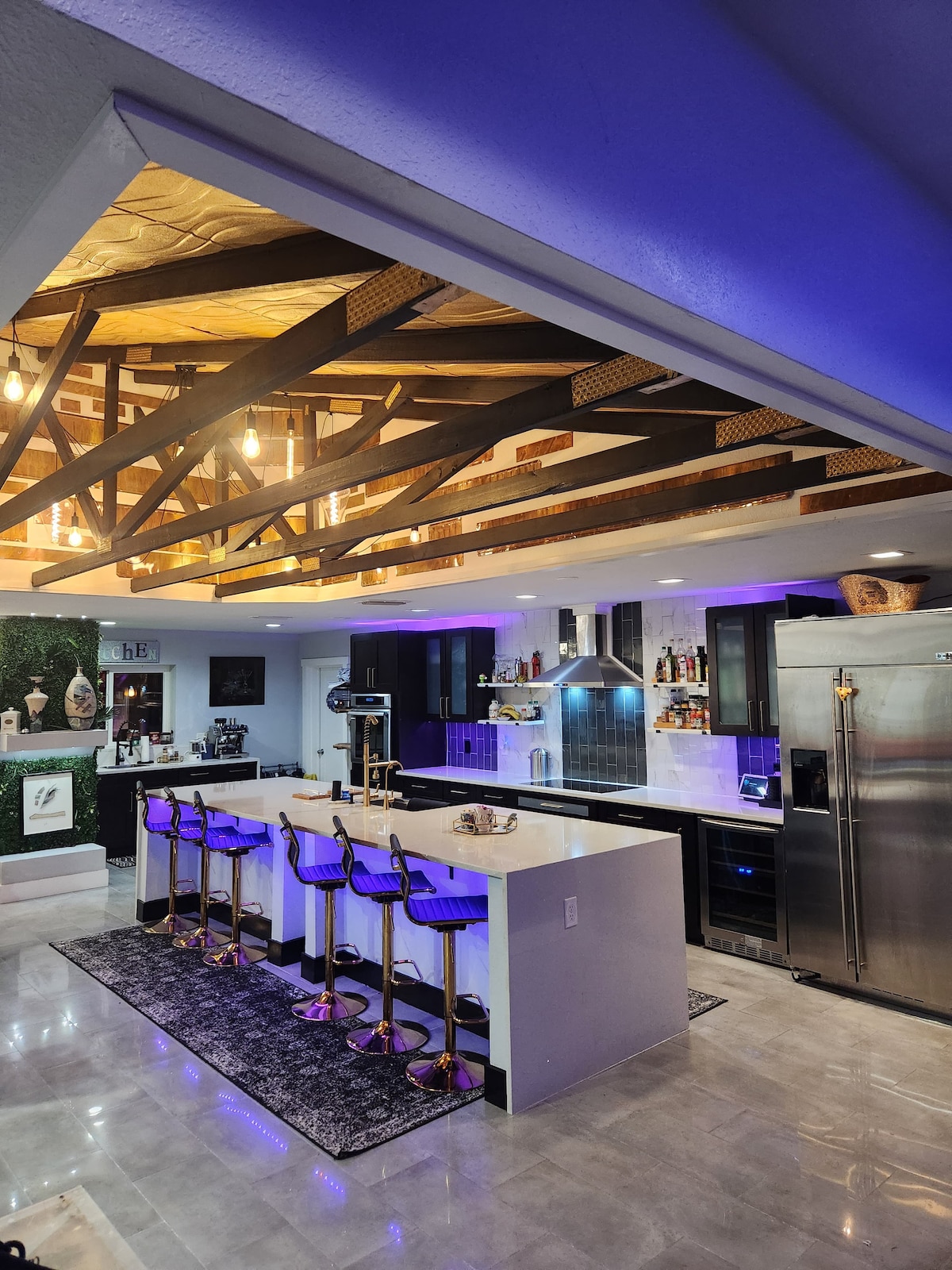 A modern kitchen layout is visible, featuring sleek black cabinetry and stainless steel appliances. A raised bar area with four purple stools is illuminated by warm pendant lighting. Exposed wooden beams add character to the ceiling, enhancing the space's stylish decor.