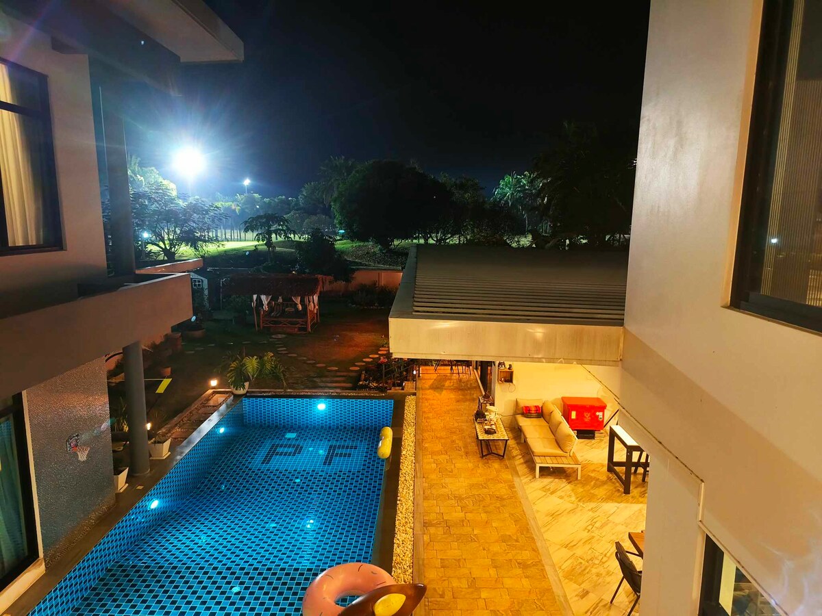 A nighttime view captures a tranquil pool surrounded by lush greenery. Soft lighting highlights the outdoor seating area, while the glow from floodlights creates a serene atmosphere. The pool's tiled surface reflects the surrounding landscape, emphasizing the feeling of relaxation.
