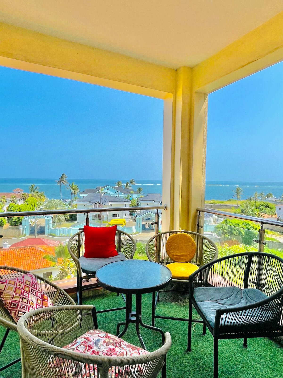 A spacious balcony features a round table surrounded by four chairs, offering a vibrant pop of color. The sea view in the background provides a serene atmosphere, with rooftops and palm trees visible below under a clear blue sky.