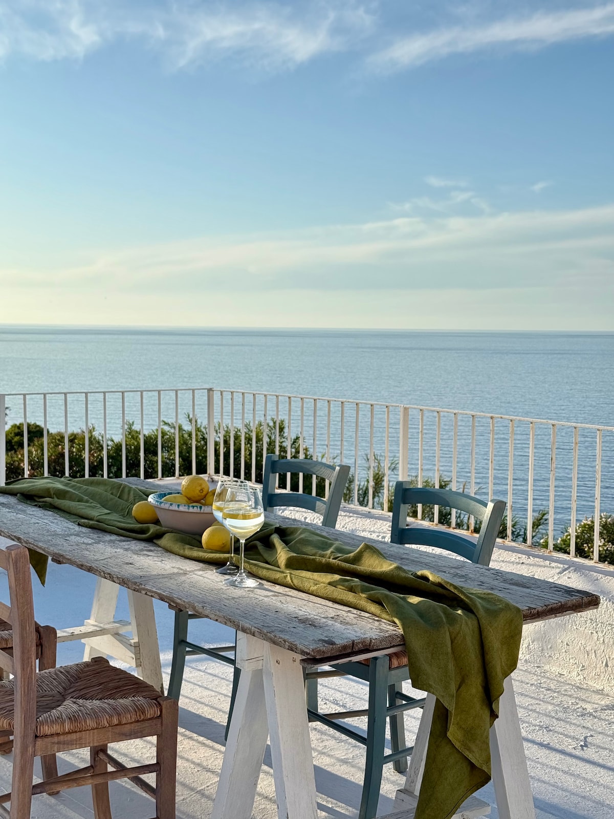 An outdoor dining space is presented with a rustic table adorned with a green cloth and a bowl of lemons. Glasses filled with a light drink sit ready for enjoyment, facing a serene view of the blue sea and sky, framed by a white railing.