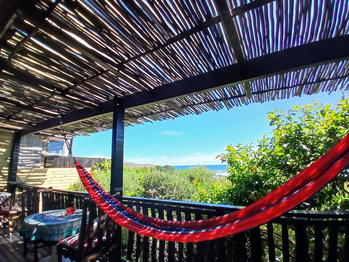 A shaded outdoor area features a vibrant red hammock suspended between wooden beams. Lush greenery surrounds the space, with a glimpse of the ocean visible in the distance. A small table and chairs are arranged, inviting relaxation and enjoyment of the scenic views.