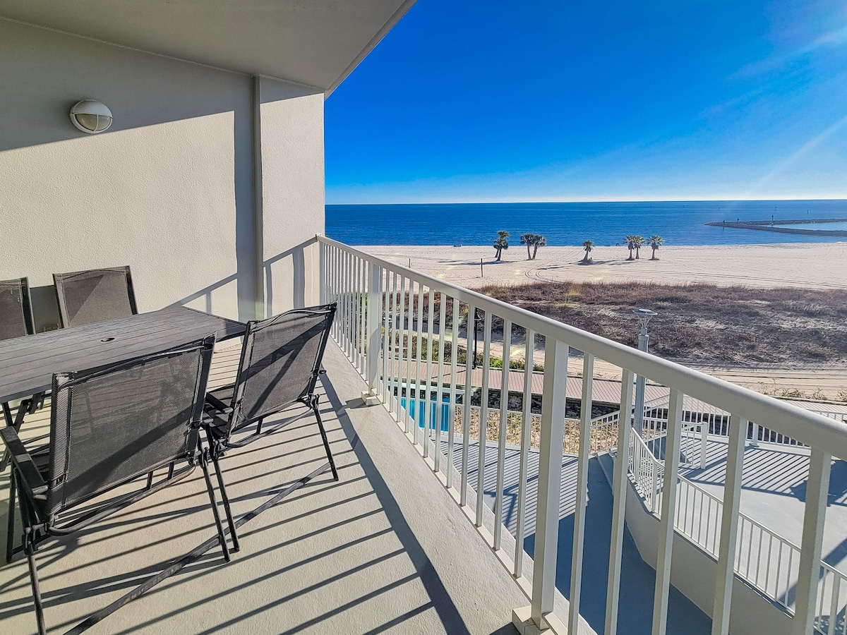 A private balcony offers expansive views of the ocean and beach, with a dining table surrounded by chairs. Sunlight casts gentle shadows across the surface, enhancing the inviting space. The beach is visible in the distance, framed by a clear sky.