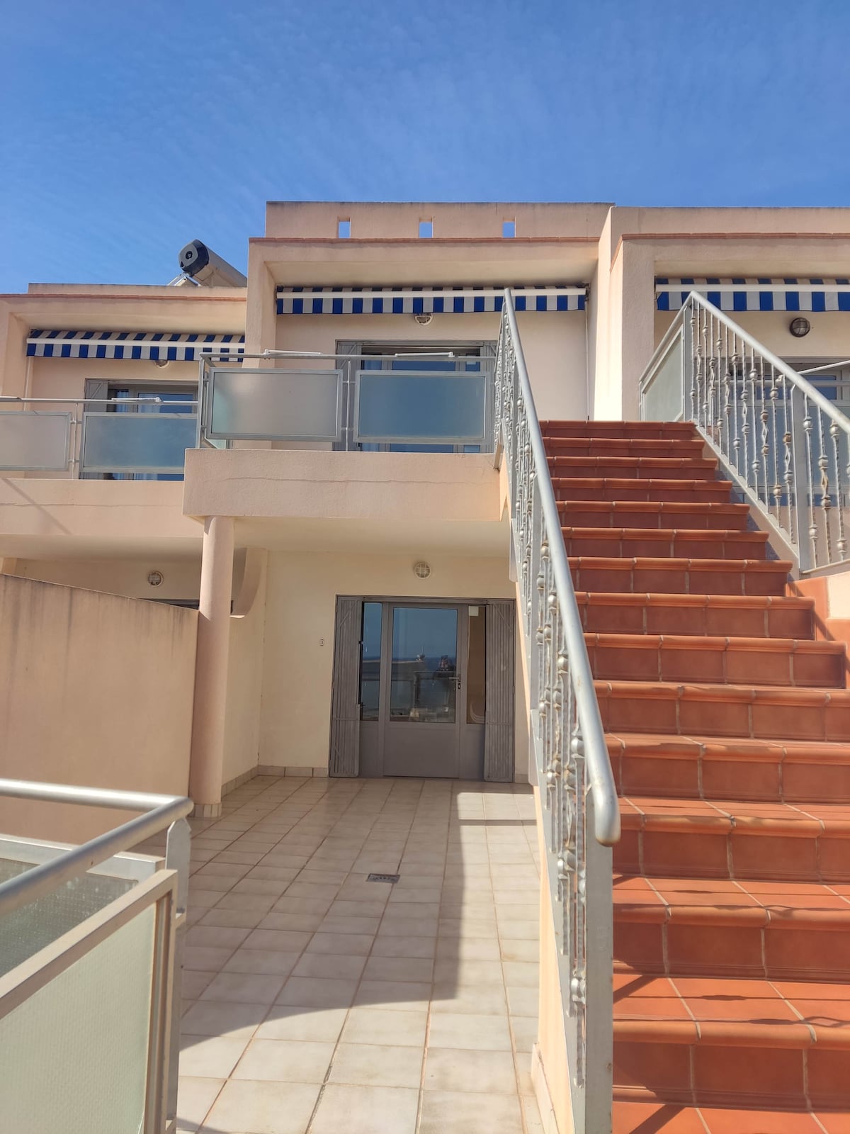An outdoor view features a staircase leading to an upper level, flanked by a railing. The facade is light-colored with striped awnings visible above the glass doors. A tiled area is seen at the base of the stairs, and a clear blue sky fills the background.